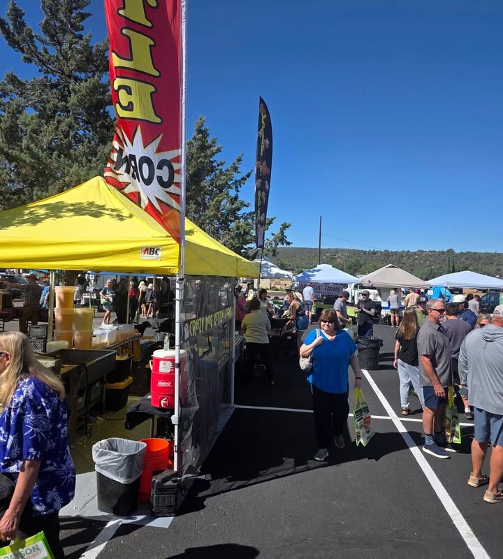 Weekend farmers market with yellow food vendor tent, shoppers browsing stalls under clear blue sky